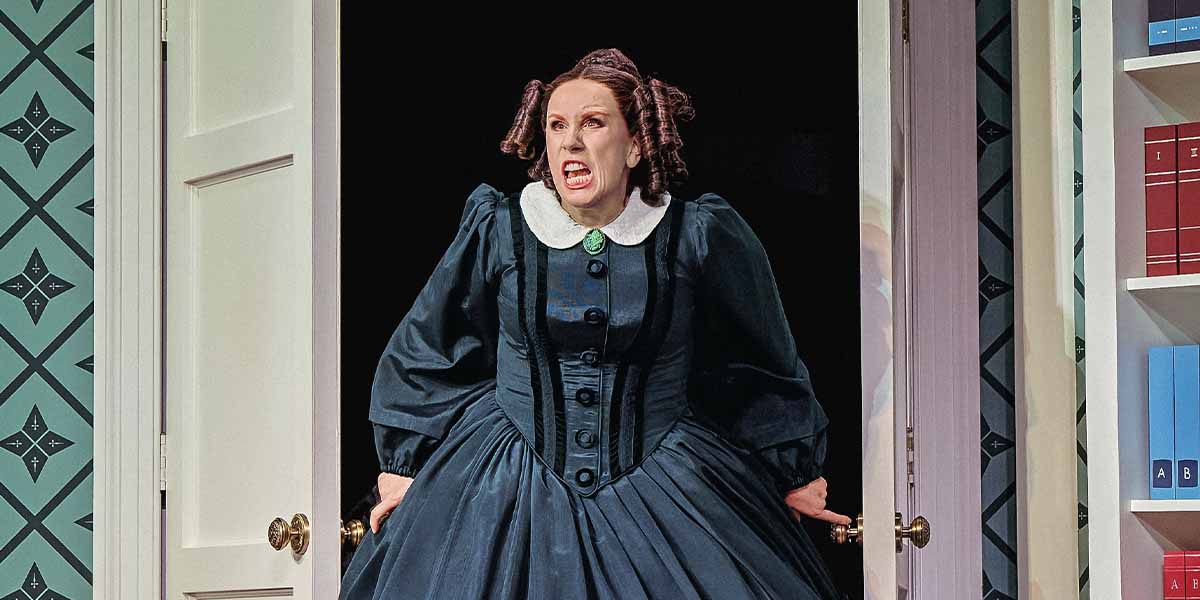 A person in a dark dress with a white collar expresses anger while standing in a doorway.