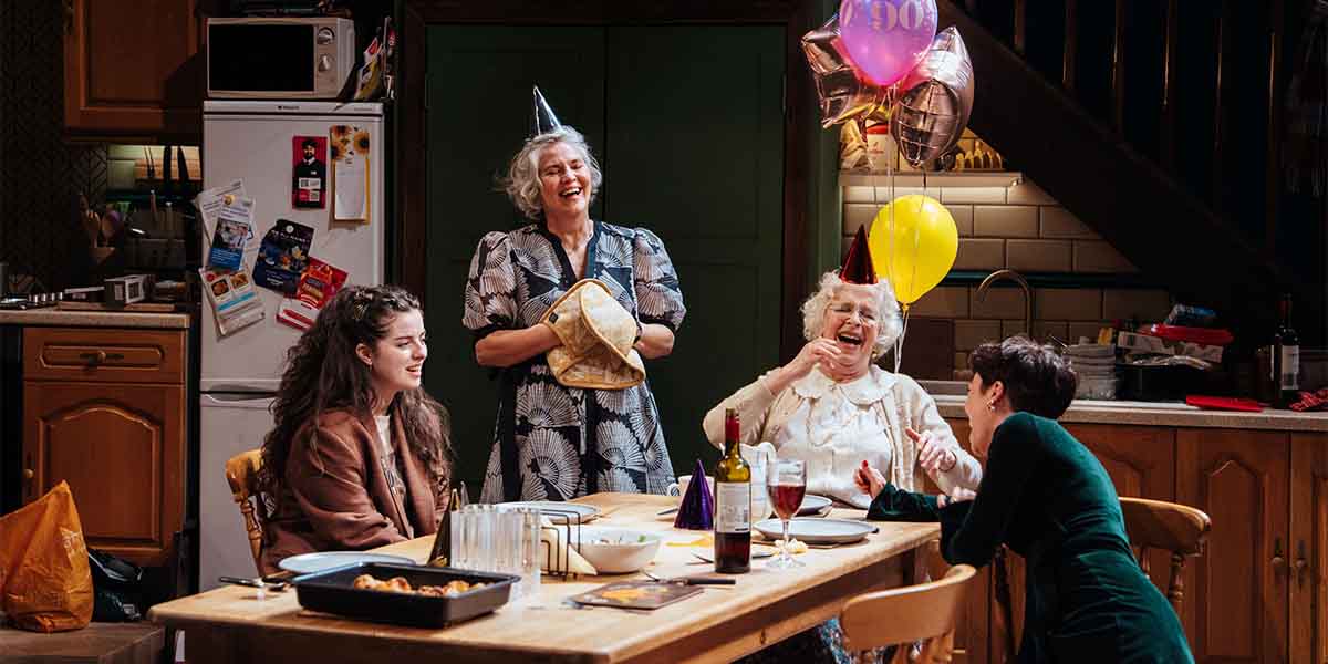 Four people celebrate around a table, wearing party hats, with balloons, food, and drinks visible.