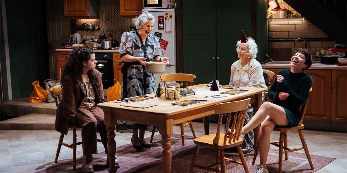 Four people sit around a wooden table, laughing; one serves food from a pot, wearing a patterned dress.