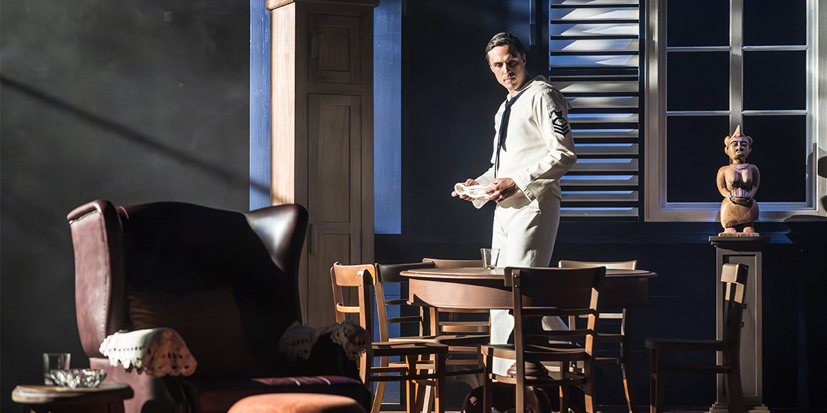 Ziggy Heath as Tom stands on stage, wearing a dark suit, with dramatic lighting and a wooden backdrop.