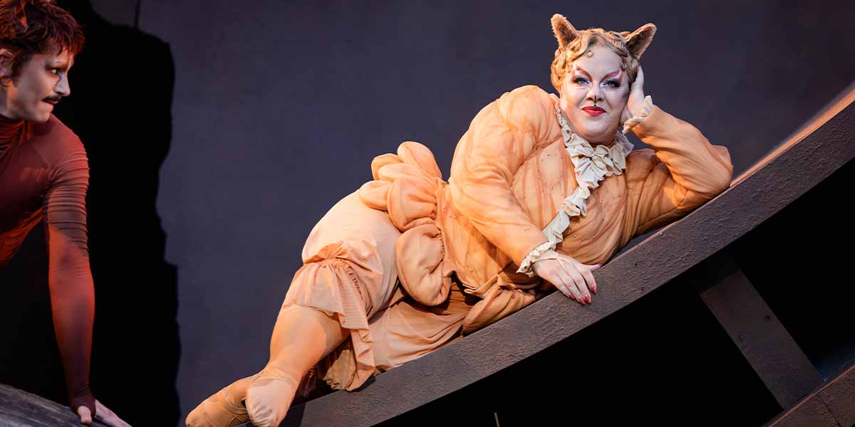 Charlotte Riby as Jennyanydots reclines on a curved surface, wearing an orange costume with cat ears.