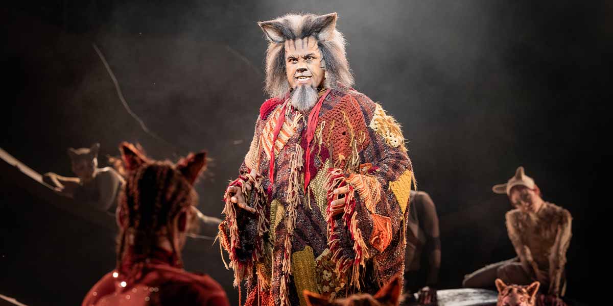Gary Wilmot as Gus, wearing a colourful, textured costume, performs on stage with other actors in the background.