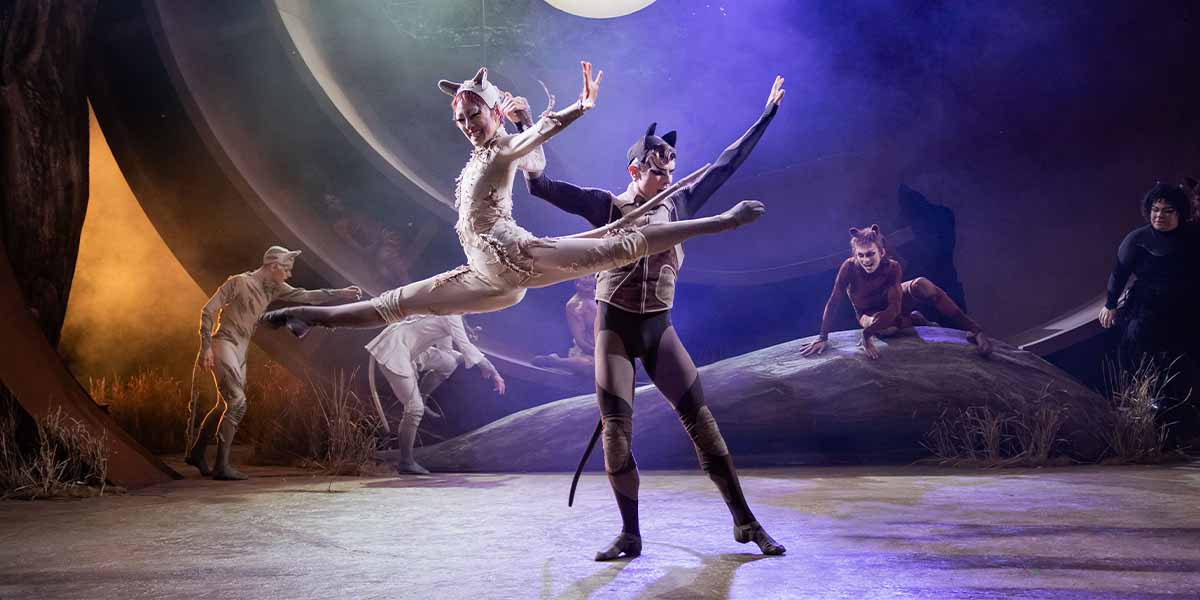 Patricia Zhou as Cassandra performs a leap with Jack Wilcox as Munkustrap; cast members in background.