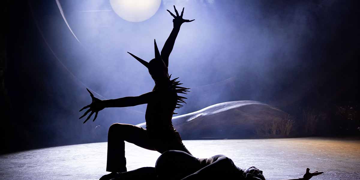 A silhouetted performer with spiked costume poses dramatically against a misty backdrop, illuminated by soft light.