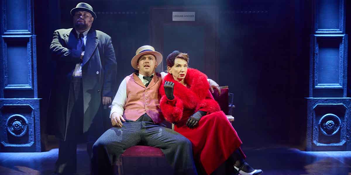 Three performers on stage sitting on a red chair.