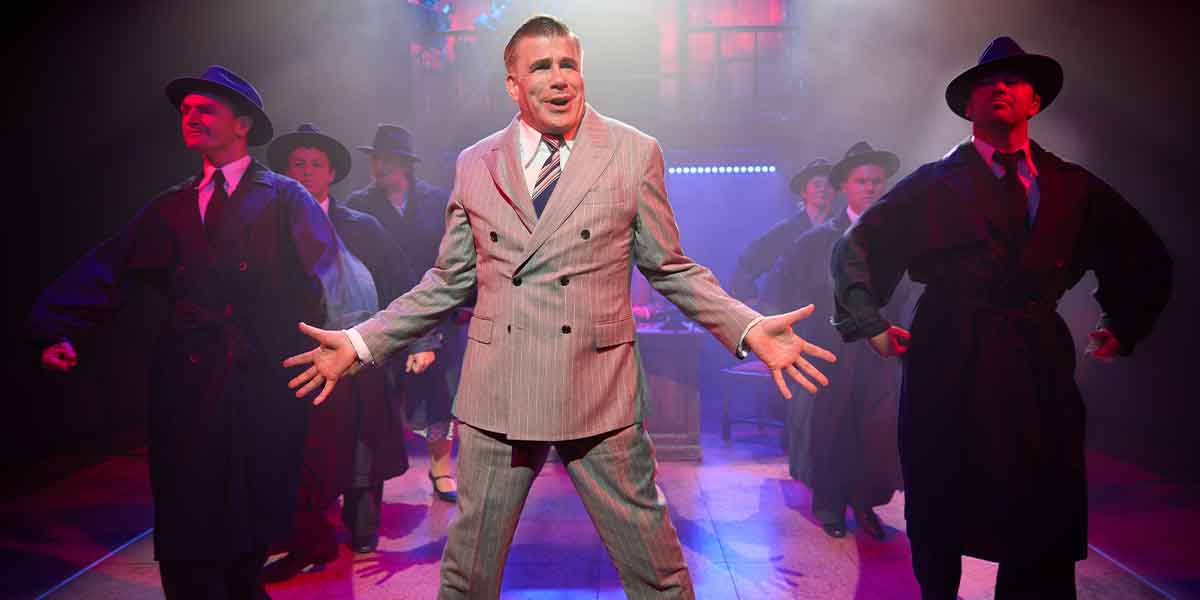 Bryan Batt performs centre stage in a grey suit, surrounded by dancers in dark coats and hats.