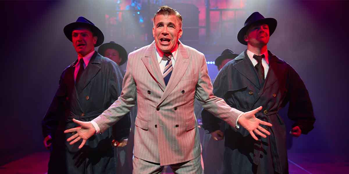 Jeremy Batt, in a striped suit, performs passionately with three men in dark coats and hats.