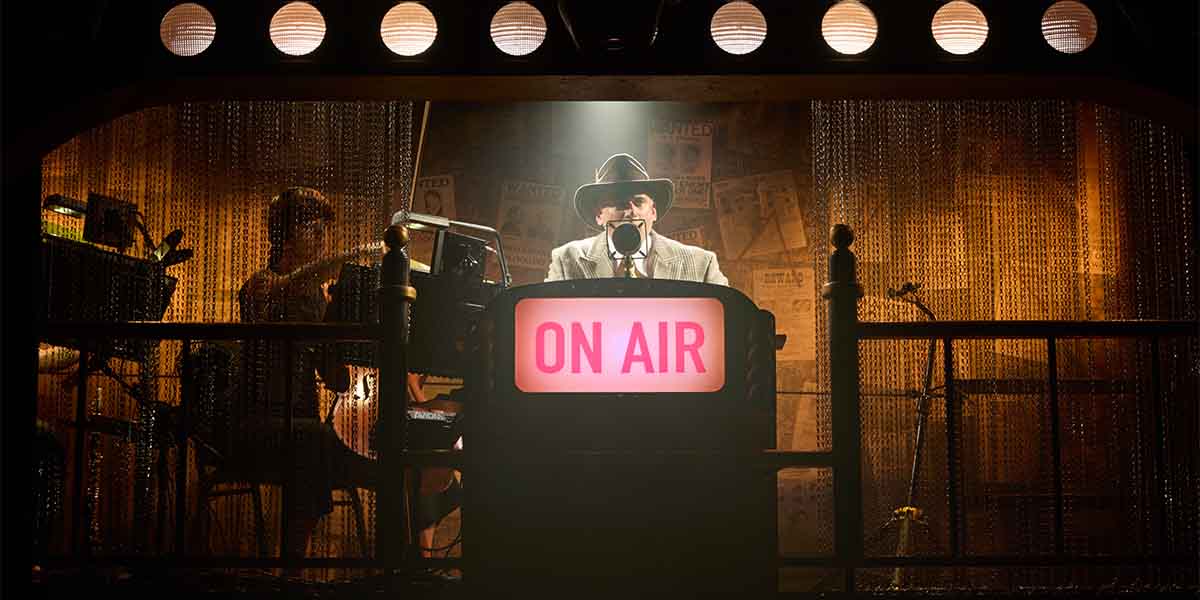 Marc Elliott, dressed in a light suit and hat, speaks into a microphone at an "ON AIR" desk.