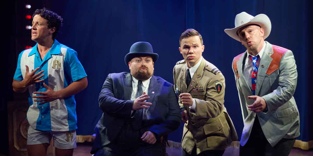 Four performers in theatrical costumes expressively pose on stage, with a blue backdrop and dramatic lighting.