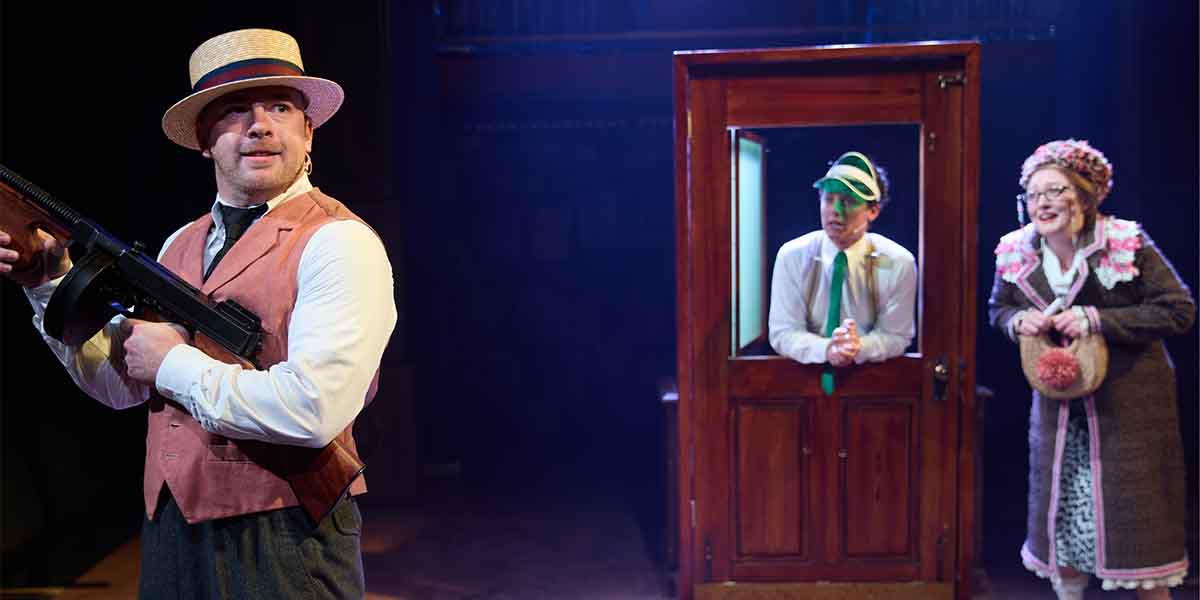 A person in a straw hat holds a gun, while two others watch from a doorway, dressed in period costumes.