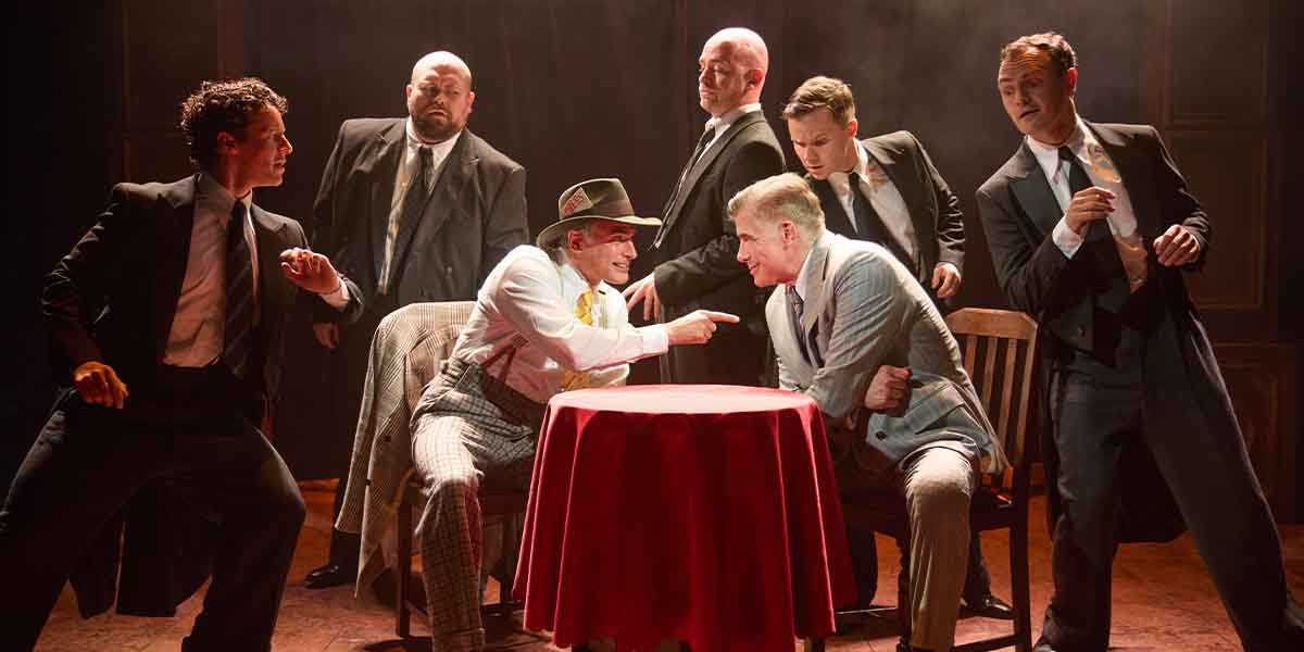 A group of seven people in formal attire interact around a red tablecloth, with dramatic lighting.
