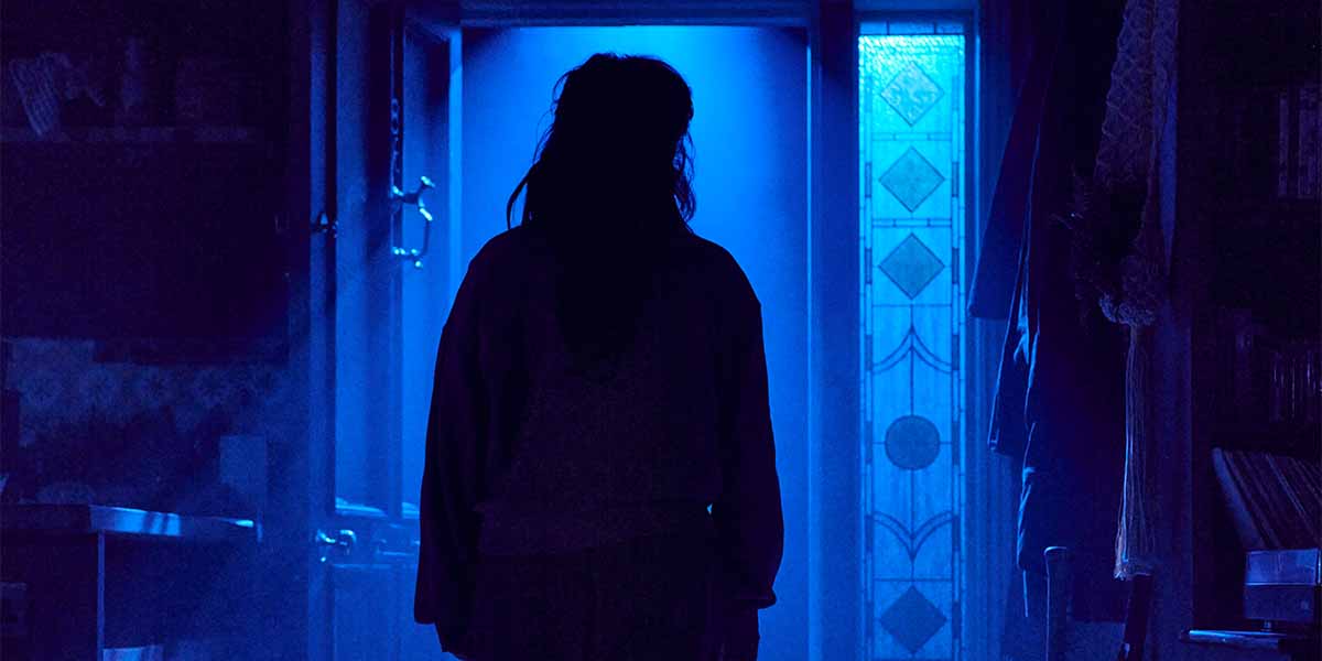 Ami Okumura Jones as 'Lou' stands silhouetted against a blue-lit doorway, surrounded by shadows.