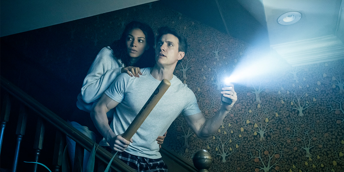 two people stand in a dimly lit room, looking intently at an eerie, shadowy figure.