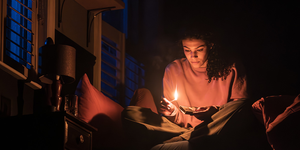 A person sits cross-legged on a bed, holding a lit candle, with a focused expression in low light.