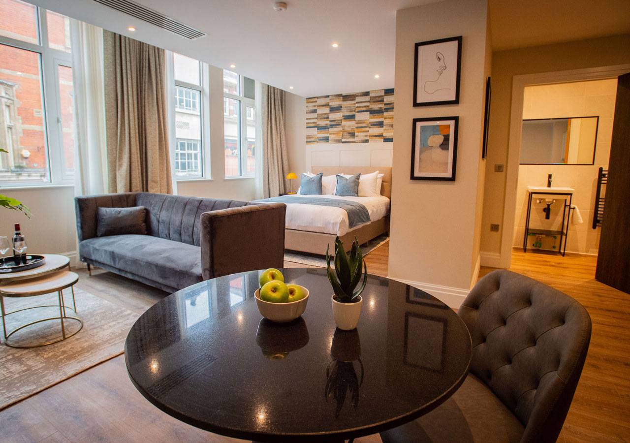 A modern hotel studio featuring a double bed, wooden furniture, and large windows with natural light.