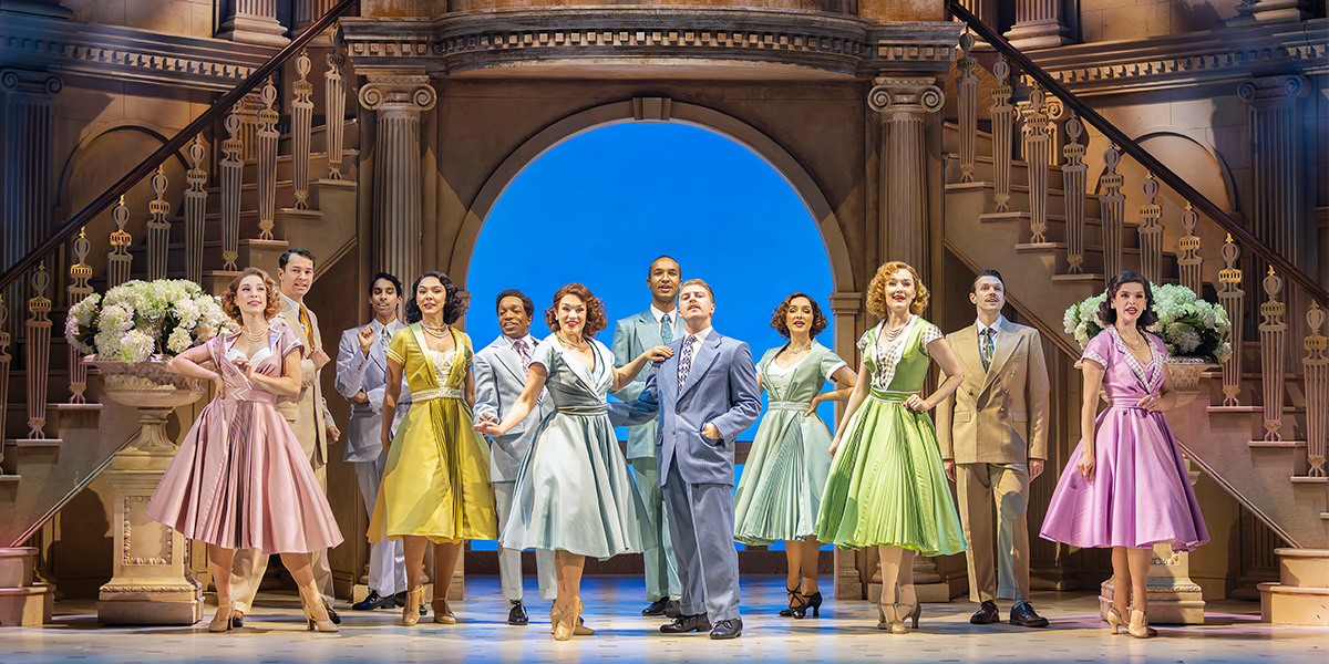 The cast of High Society poses together, dressed in elegant attire, against a soft, neutral background.