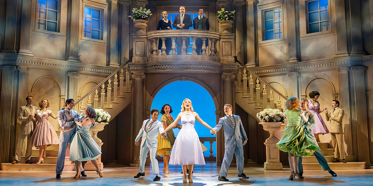 Helen George as Tracy Lord stands centre, surrounded by cast members in elegant attire, smiling at the camera.