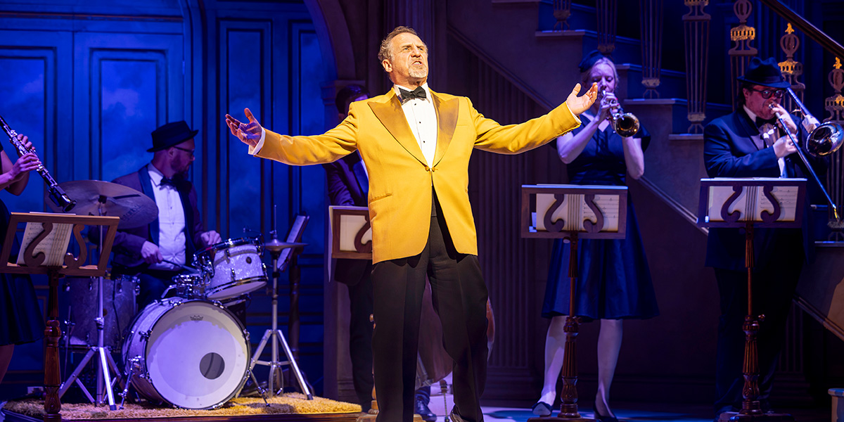 Nigel Lindsay as Uncle Willie, wearing a suit, smiles against a blurred theatre backdrop.