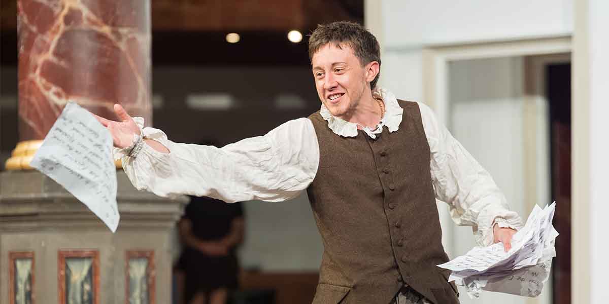 Charlie Josephine as Orlando gestures expressively, holding papers, wearing a brown vest and white shirt.