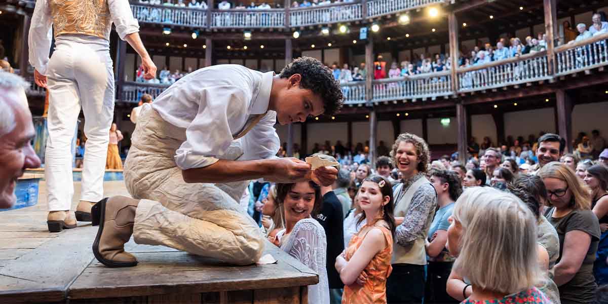 Jax Guerrero as Dumaine, dressed in period costume, performs on stage at Shakespeare's Globe Theatre.