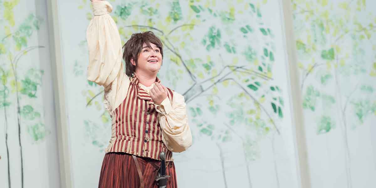 Lola Shalam as Rosalind, smiling and raising her hand, wearing a striped vest against a painted backdrop.