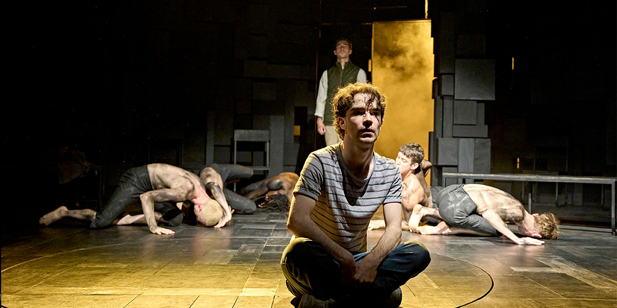 Noah Valentine as Alan Strang stands centre stage, surrounded by the cast of Equus in dramatic lighting.