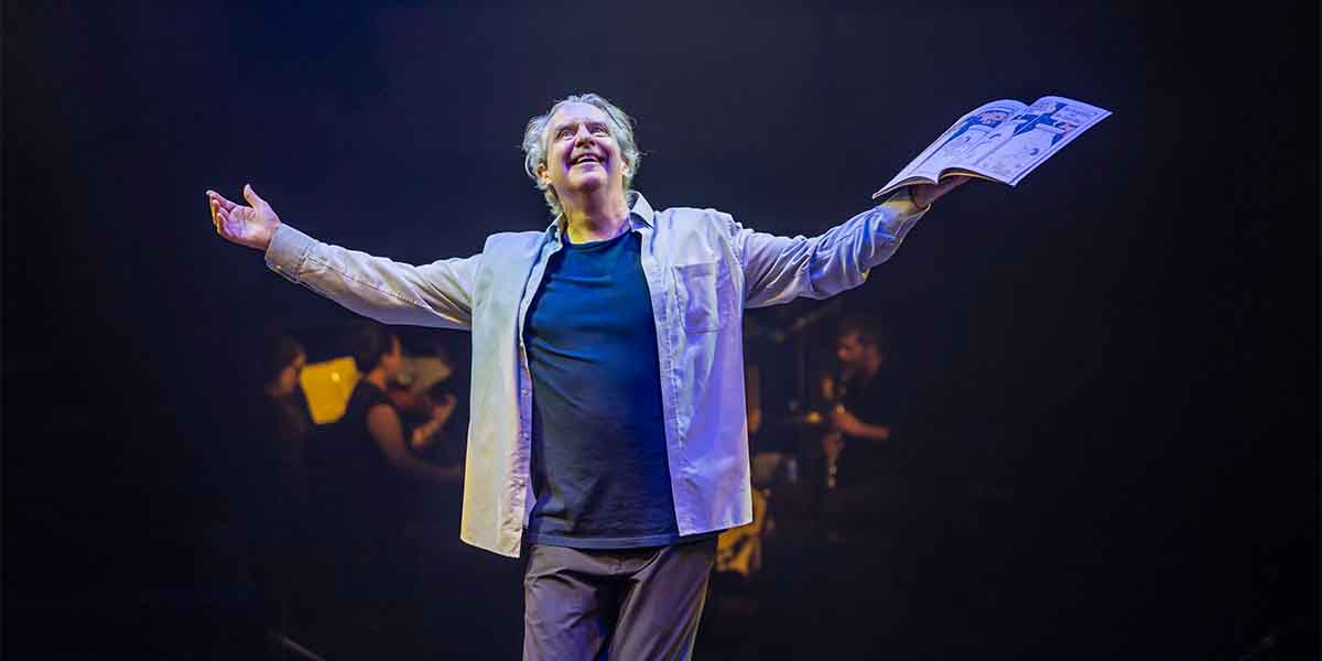 Phelim McDermott stands with arms outstretched, holding a booklet, illuminated by stage lights.