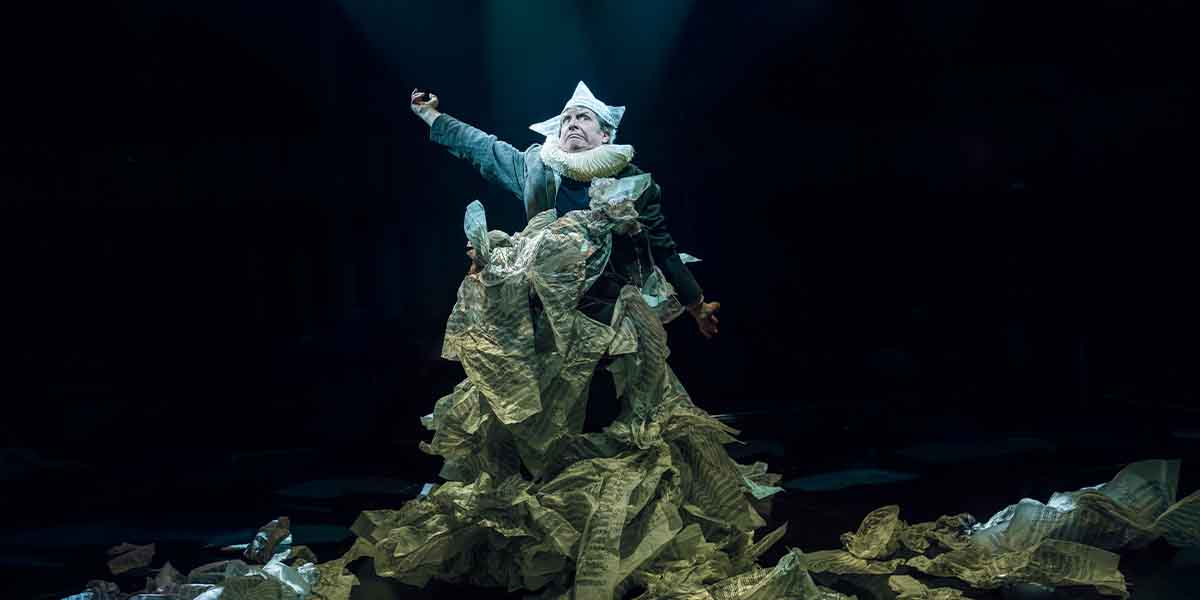 Phelim McDermott performs dramatically in a costume made of crumpled paper, under focused stage lighting.