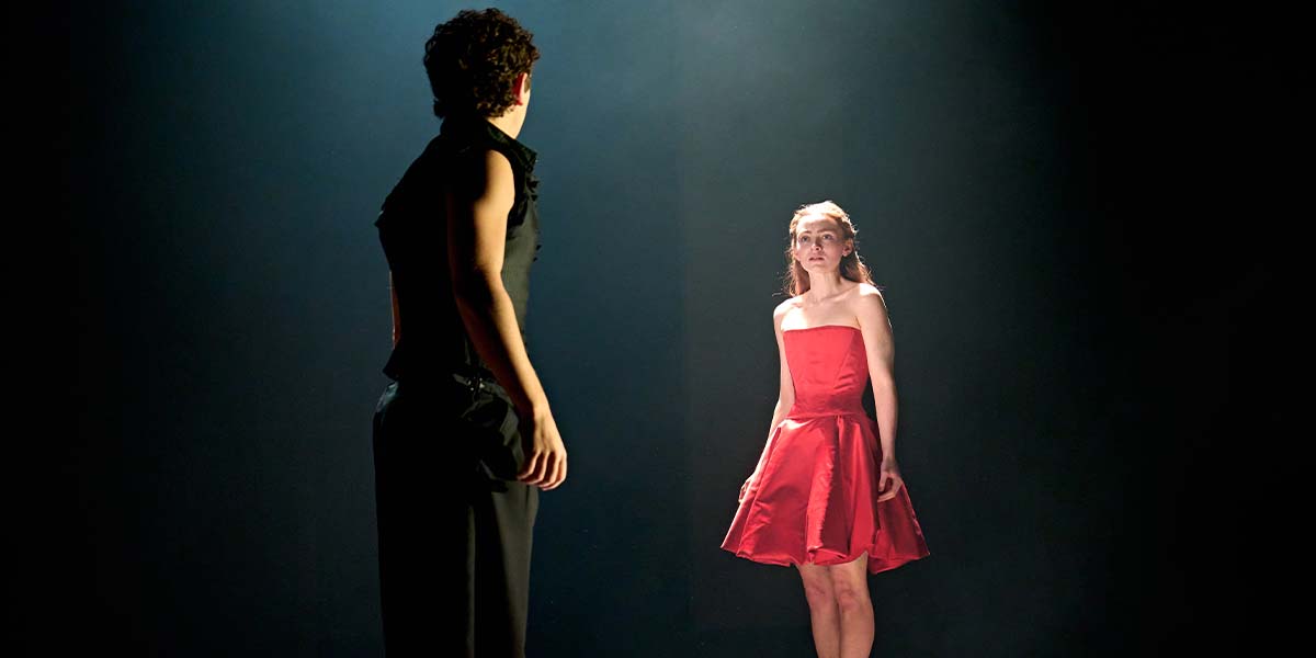 Romeo and Juliet stand facing each other, with Juliet in a red dress and dim lighting.
