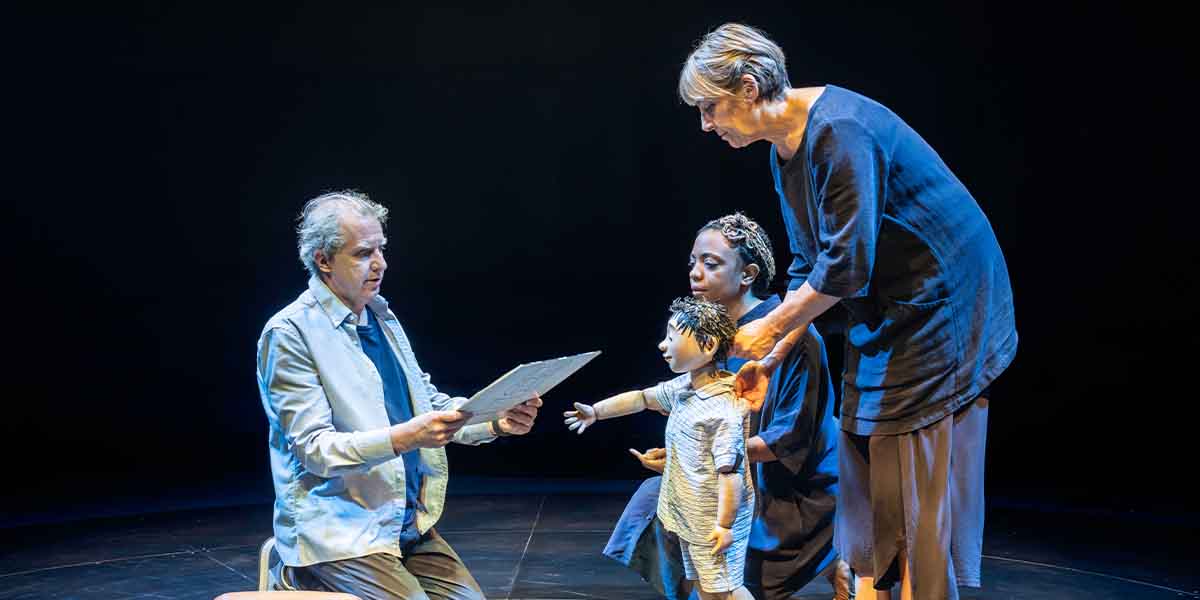 A man reads a script while two women interact with a puppet in a dark stage setting.