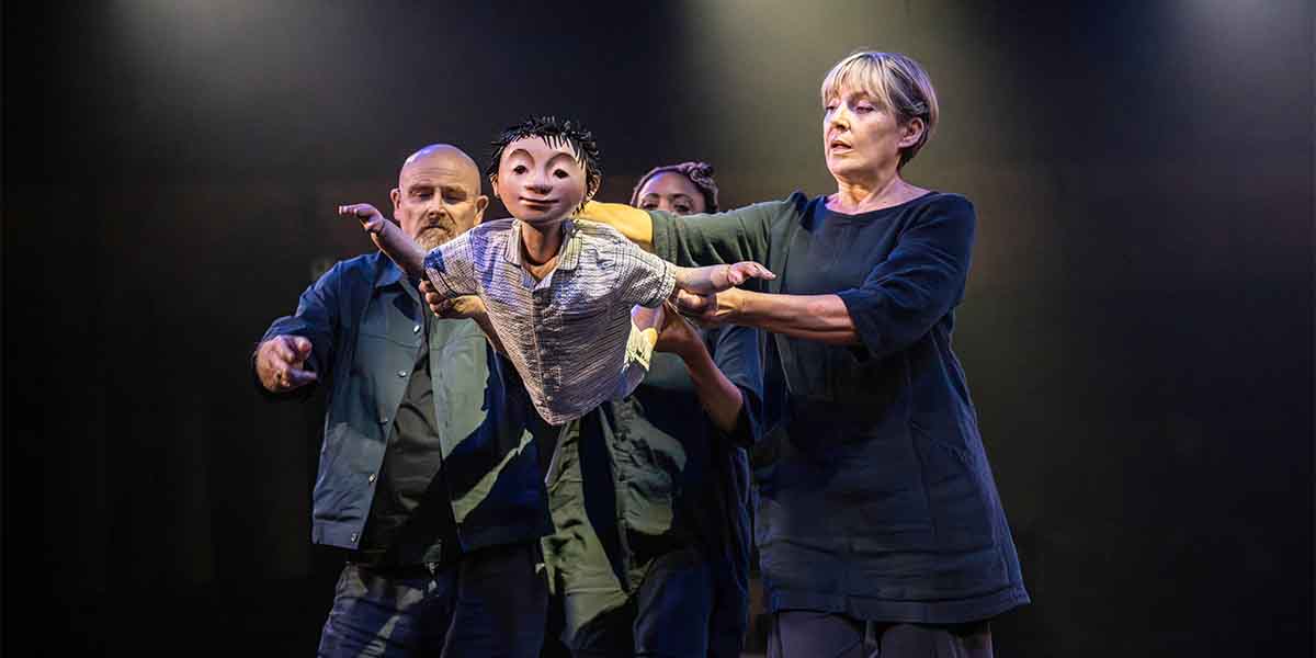 A woman in a dark outfit holds a puppet, while two others assist her on stage.