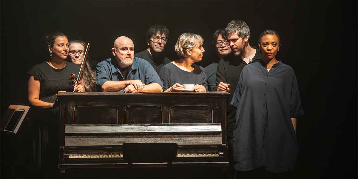 A group of eight people stands around a piano, engaged in conversation, against a dark background.