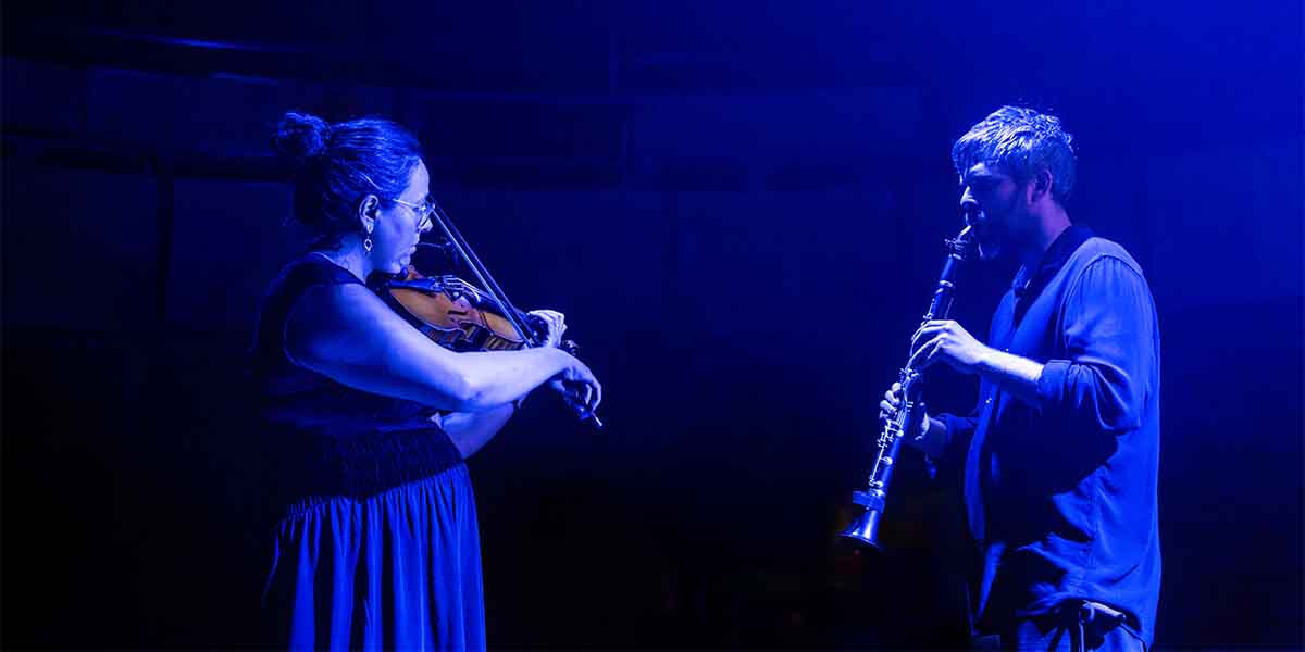 A woman plays the violin while a man plays the clarinet, both illuminated by blue lighting.