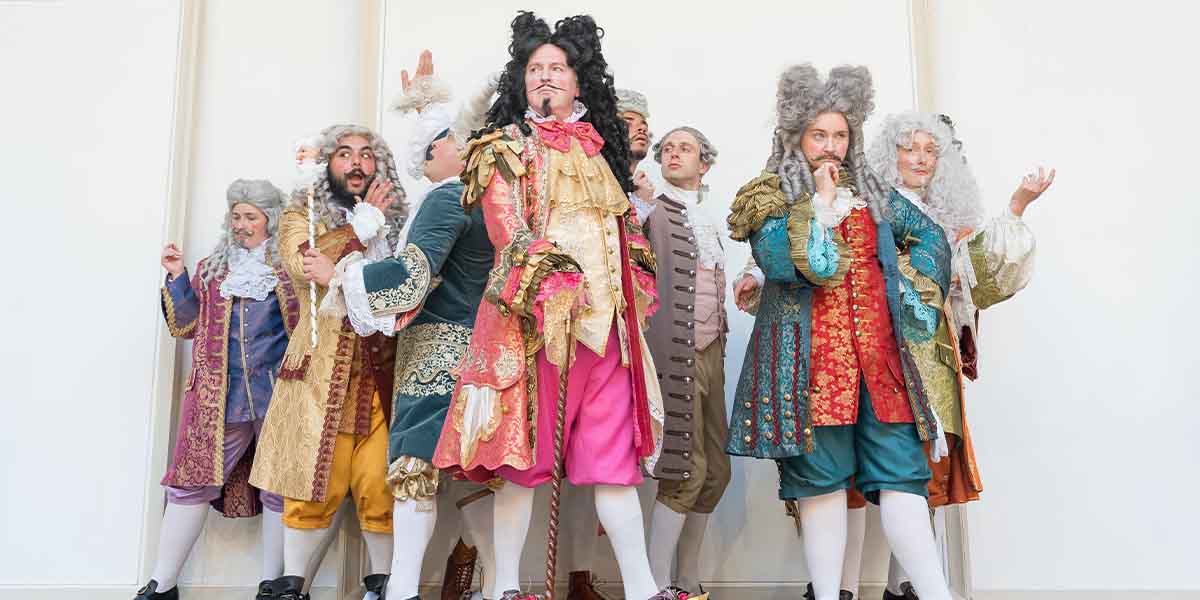 Eight actors in elaborate period costumes pose playfully, showcasing vibrant colours and intricate details.