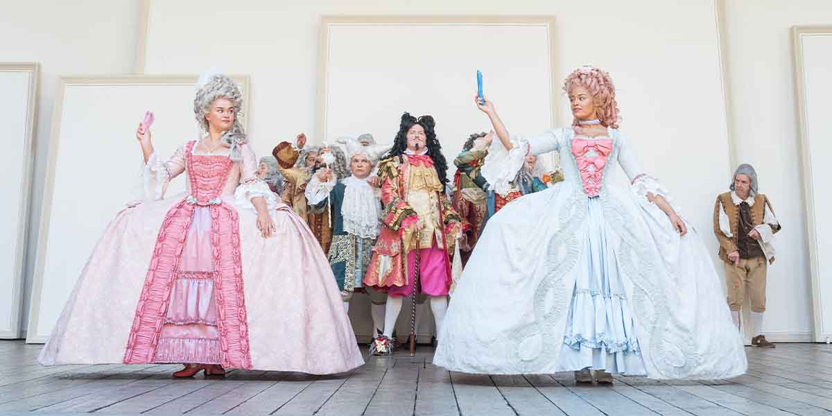 Two actresses in elaborate period costumes pose centre stage, while a group of performers stands behind them.