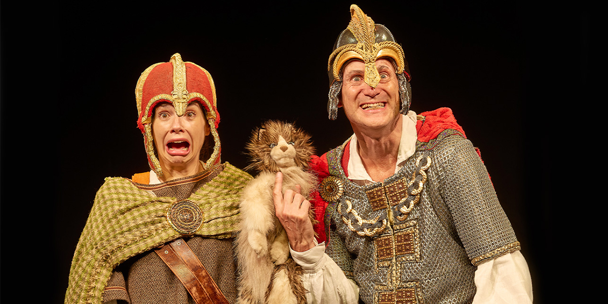Children dressed in historical costumes perform on stage during a Horrible Histories event, with vibrant scenery.