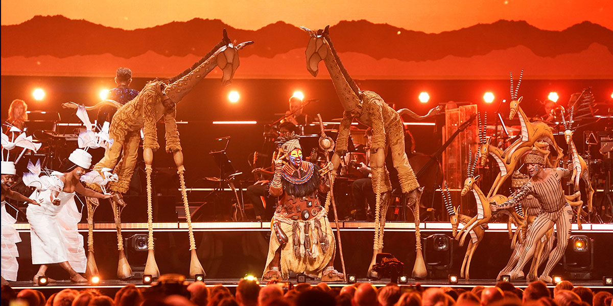 A vibrant stage scene from The Lion King featuring performers in elaborate costumes, with a jungle backdrop.