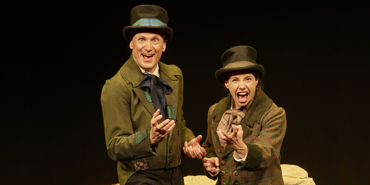 Two actors in Victorian costumes, smiling and gesturing enthusiastically against a dark background.