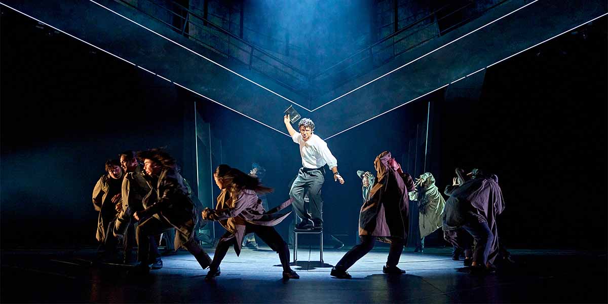 A performer in a white shirt stands on a stool, holding a book, surrounded by dancers in dark cloaks.