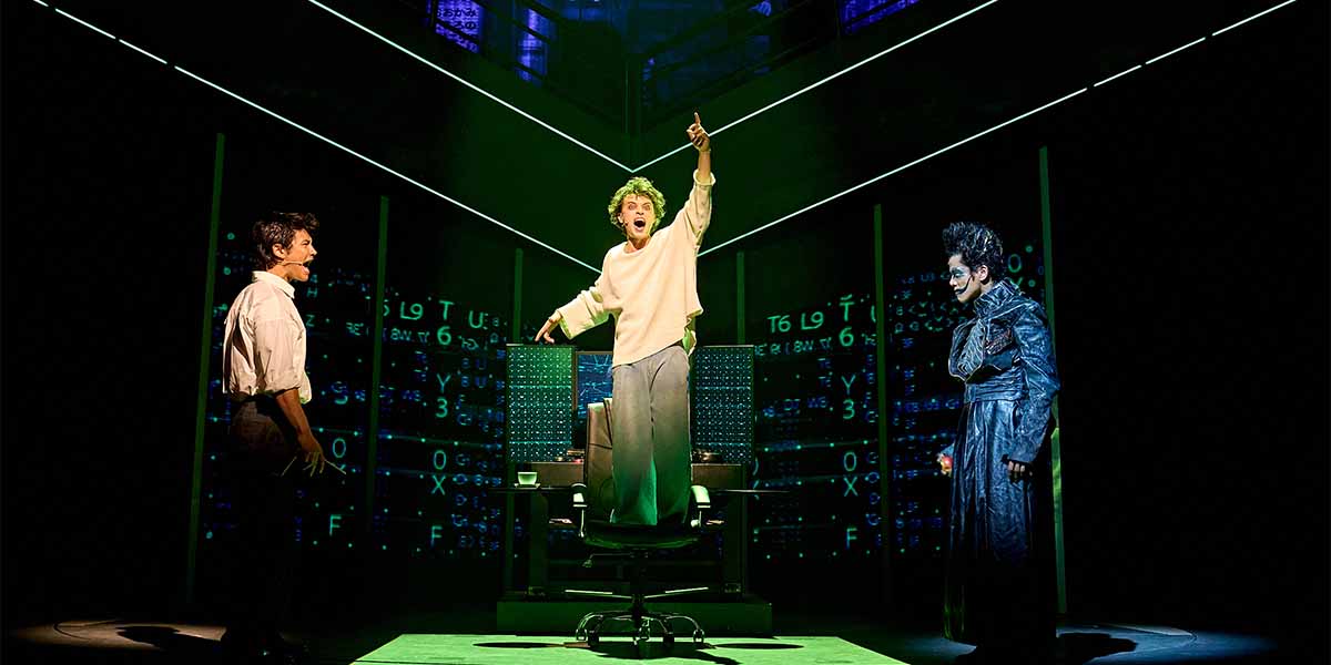 Three performers on stage: one stands excitedly in a chair, while two others observe intently.