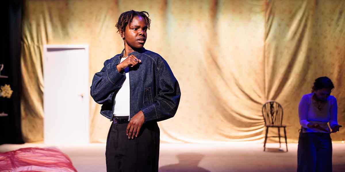 Francesca Amewudah-Rivers, dressed in a denim jacket, stands thoughtfully on stage with a blurred figure nearby.