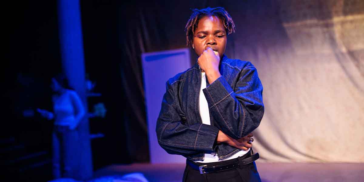 Francesca Amewudah-Rivers, in a denim jacket, ponders thoughtfully on stage, with a blurred figure behind.