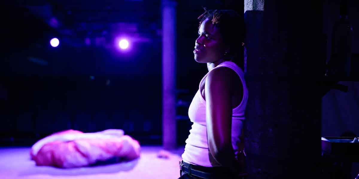 Francesca Amewudah-Rivers leans against a pillar, illuminated by purple stage lights, with a soft backdrop.