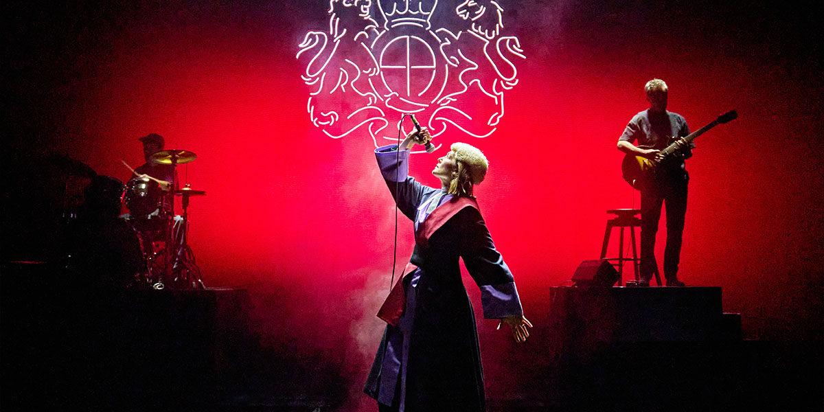 A stage scene featuring actors in period costumes performing a dramatic scene, with a richly decorated backdrop.