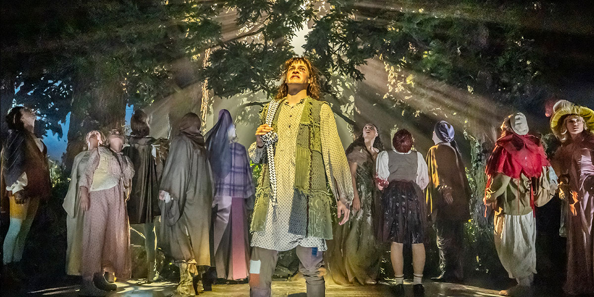 Francesca Lynn performs on stage in a forest setting, wearing a flowing dress, surrounded by trees and foliage.