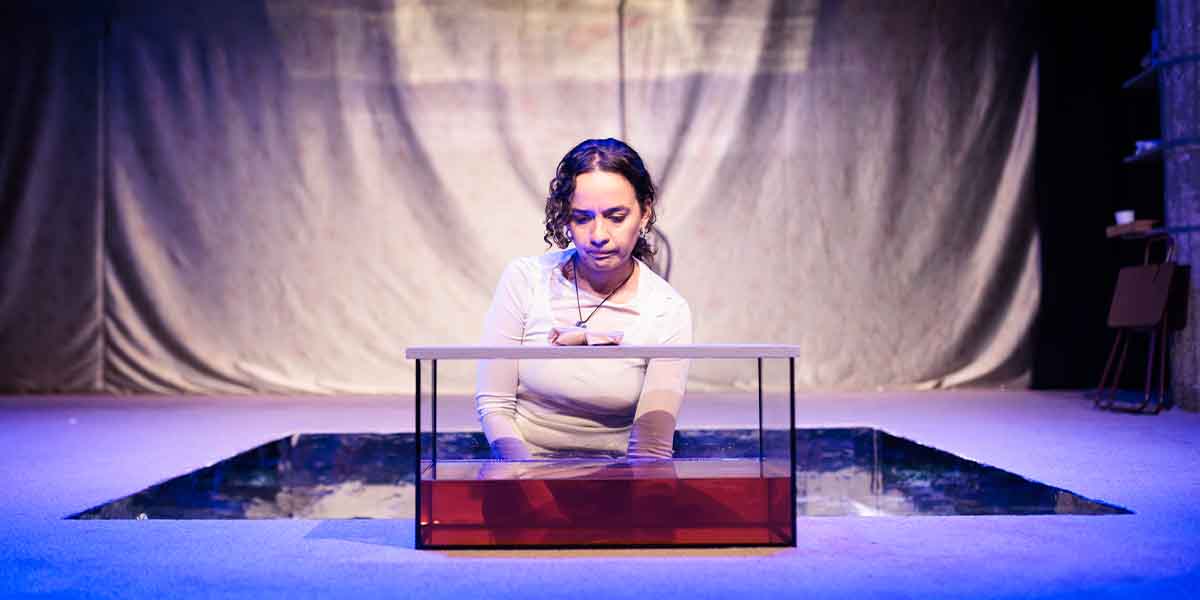 Nadi Kemp-Sayfi, as Ivy, interacts with a glass container on stage, surrounded by dim lighting.