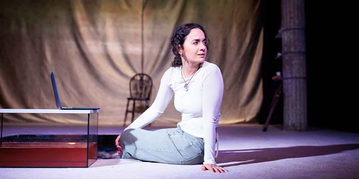 Nadi Kemp-Sayfi as Ivy sits on the floor, looking thoughtfully at a laptop, with a chair behind.
