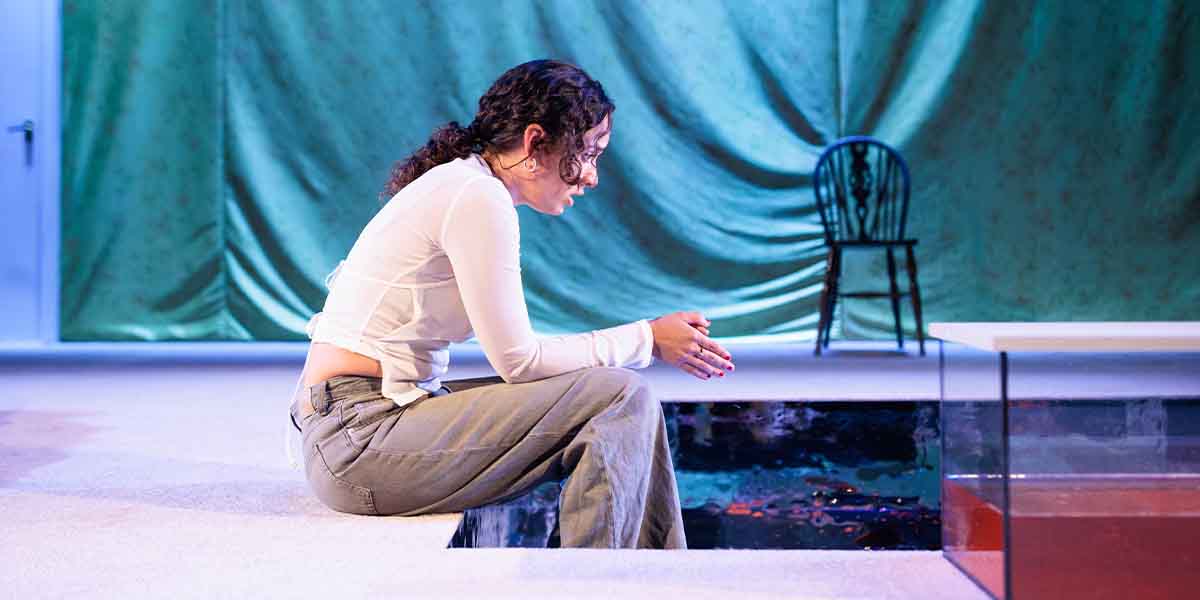 Nadi Kemp-Sayfi as Ivy sits pensively on stage, wearing a white top and grey trousers.
