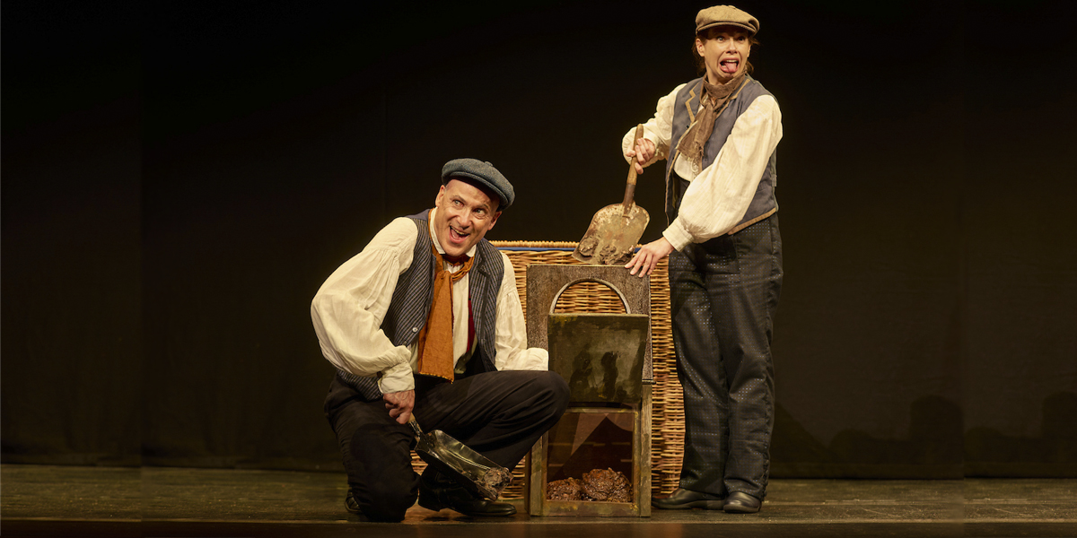 Two performers in historical costumes interact comically with a prop resembling a toilet, on stage.