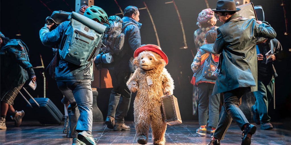 Paddington Bear in a blue coat and red hat stands centre stage, surrounded by colourful set designs and props.
